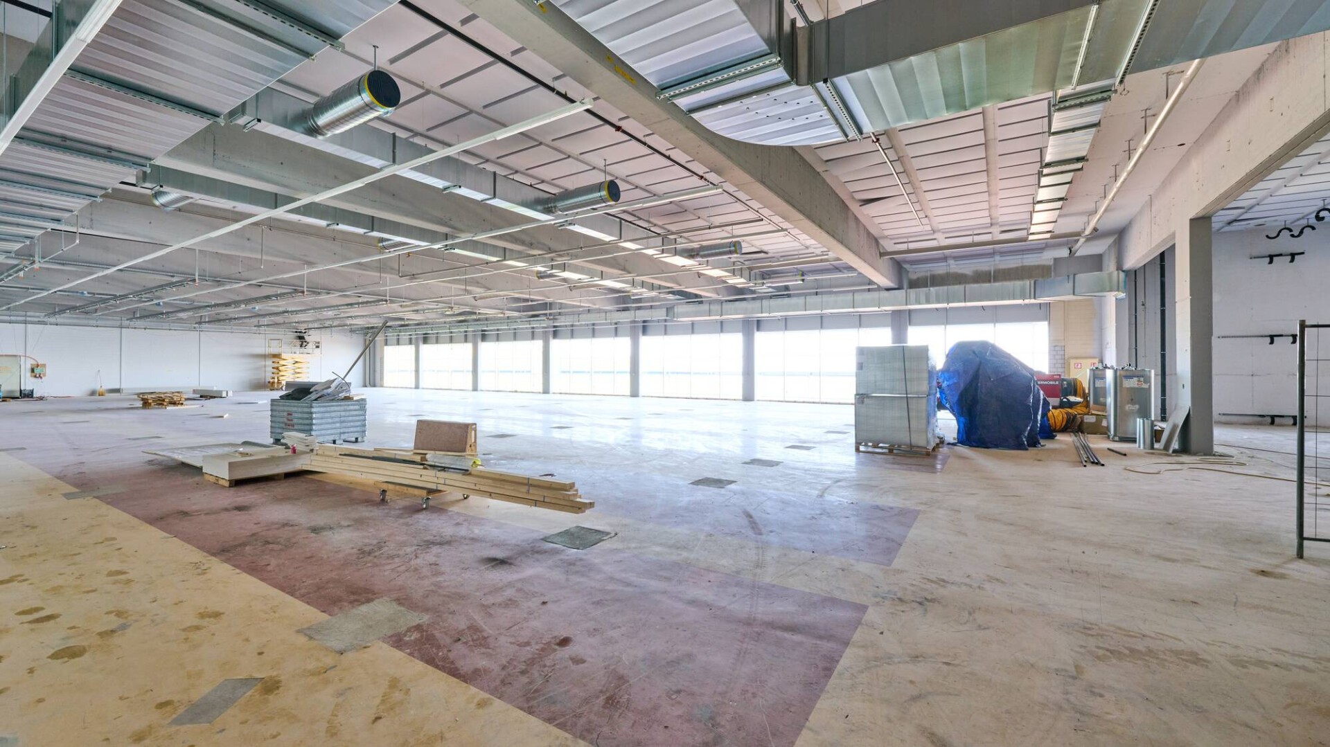 Eine große, leere Industriehalle mit freiliegenden Deckenbalken, Rohrleitungen und verstreuten Baumaterialien auf dem Boden. Sonnenlicht fällt durch breite Fenster an einer Wand ein.