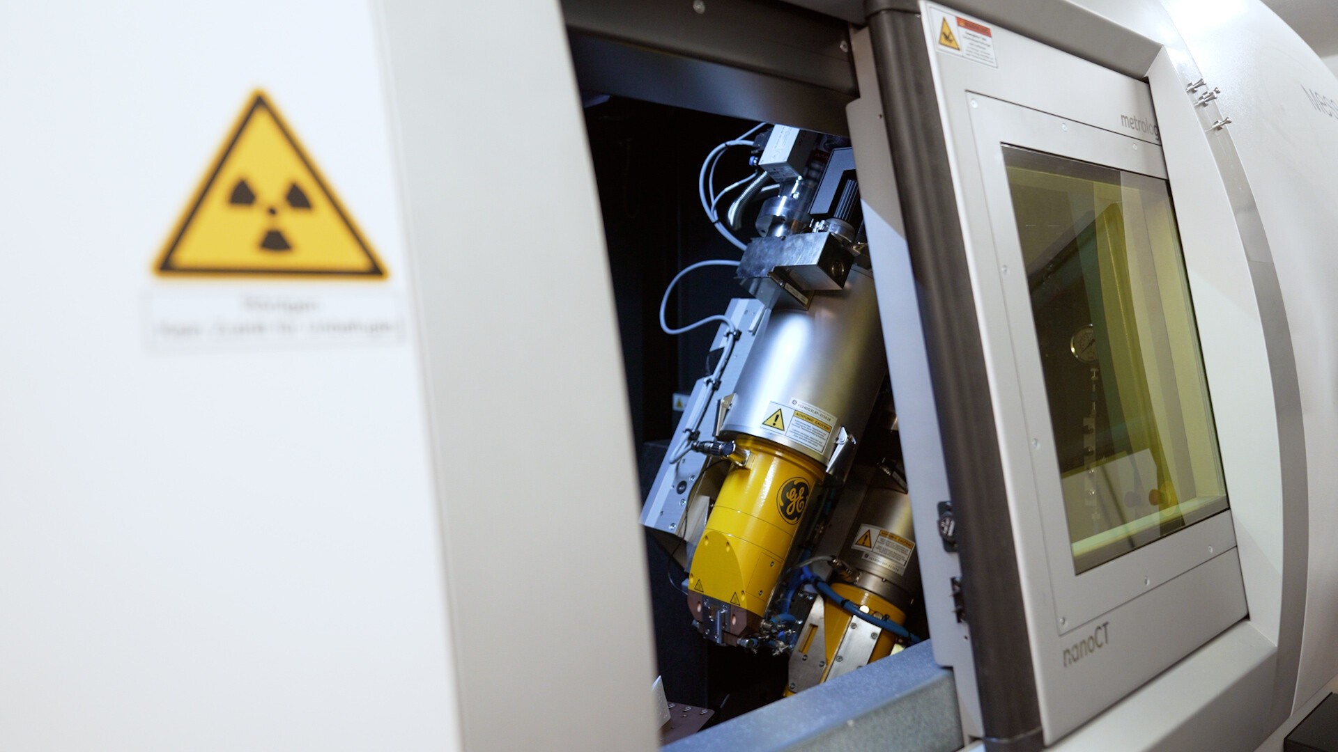 Nahaufnahme einer Industrieanlage im Inneren einer Maschine. Links ist ein Strahlungswarnschild zu sehen, im geöffneten Bedienfeld sind mechanische Komponenten und Verkabelung zu sehen. Rechts sind ein Monitor und ein Bedienfeld zu sehen.