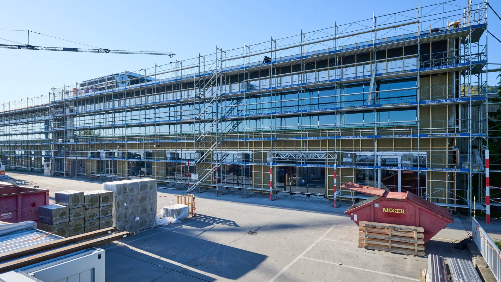 Ein modernes, mehrstöckiges Gebäude im Bau, umgeben von Gerüsten. Baumaterialien und Geräte sind über die gesamte Baustelle verstreut, und im Hintergrund ist unter einem strahlend blauen Himmel ein Kran zu sehen.