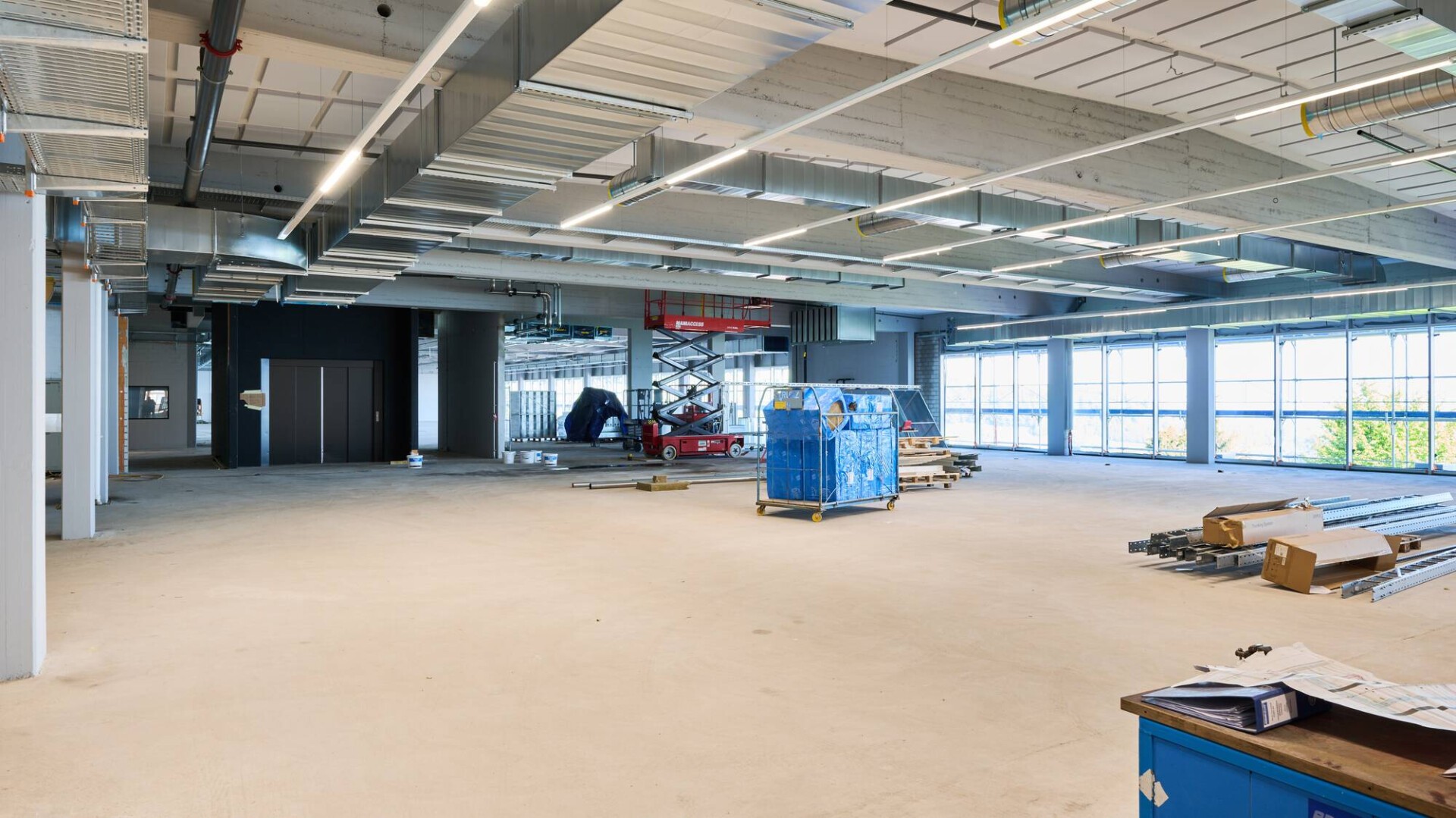 Ein geräumiges, unfertiges Büro mit freiliegenden Decken, Leitungen, Baumaterialien und großen Fenstern, die natürliches Licht hereinlassen. Geräte und Vorräte liegen verstreut auf dem nackten Boden.