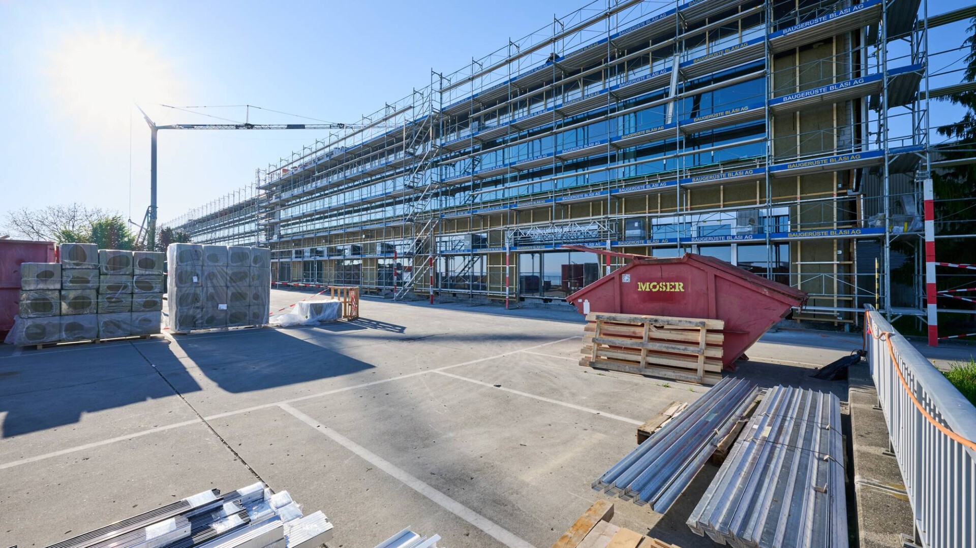 Ein modernes Gebäude im Bau mit Gerüst, Glaspaneelen und Isolierung. Baumaterialien, Paletten und ein roter Müllcontainer stehen unter einem klaren, sonnigen Himmel auf dem Parkplatz.