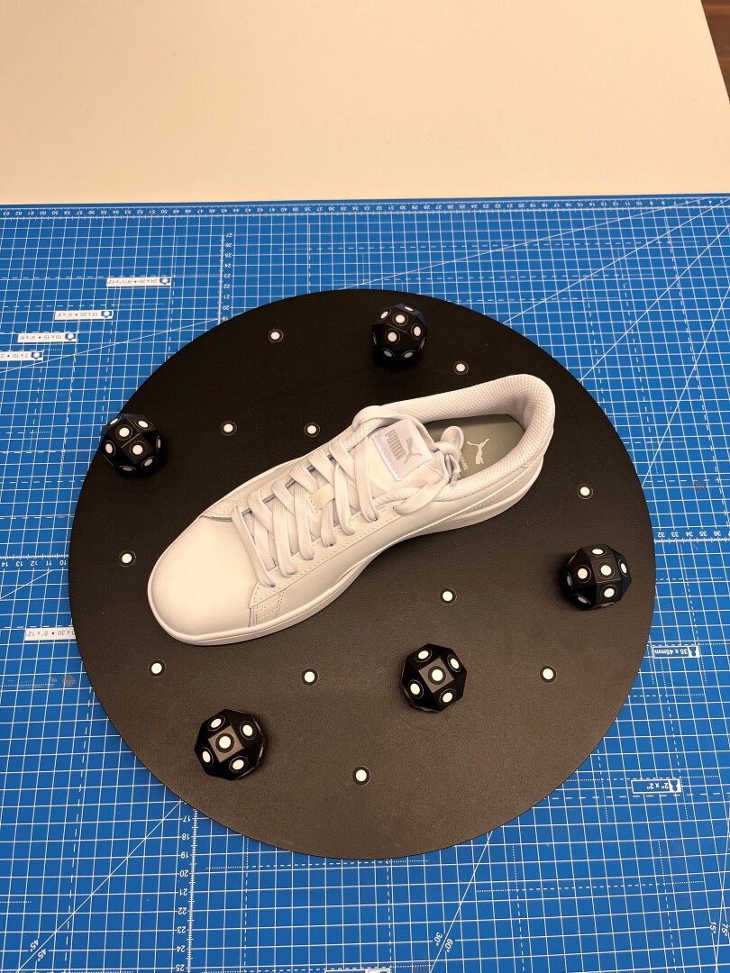 Ein weißer Turnschuh steht auf einem schwarzen runden Brett mit kleinen weißen Stiften, umgeben von fünf schwarzen 12-seitigen Würfeln mit weißen Zahlen, alles auf einer blauen Schneidematte mit Gitternetz.