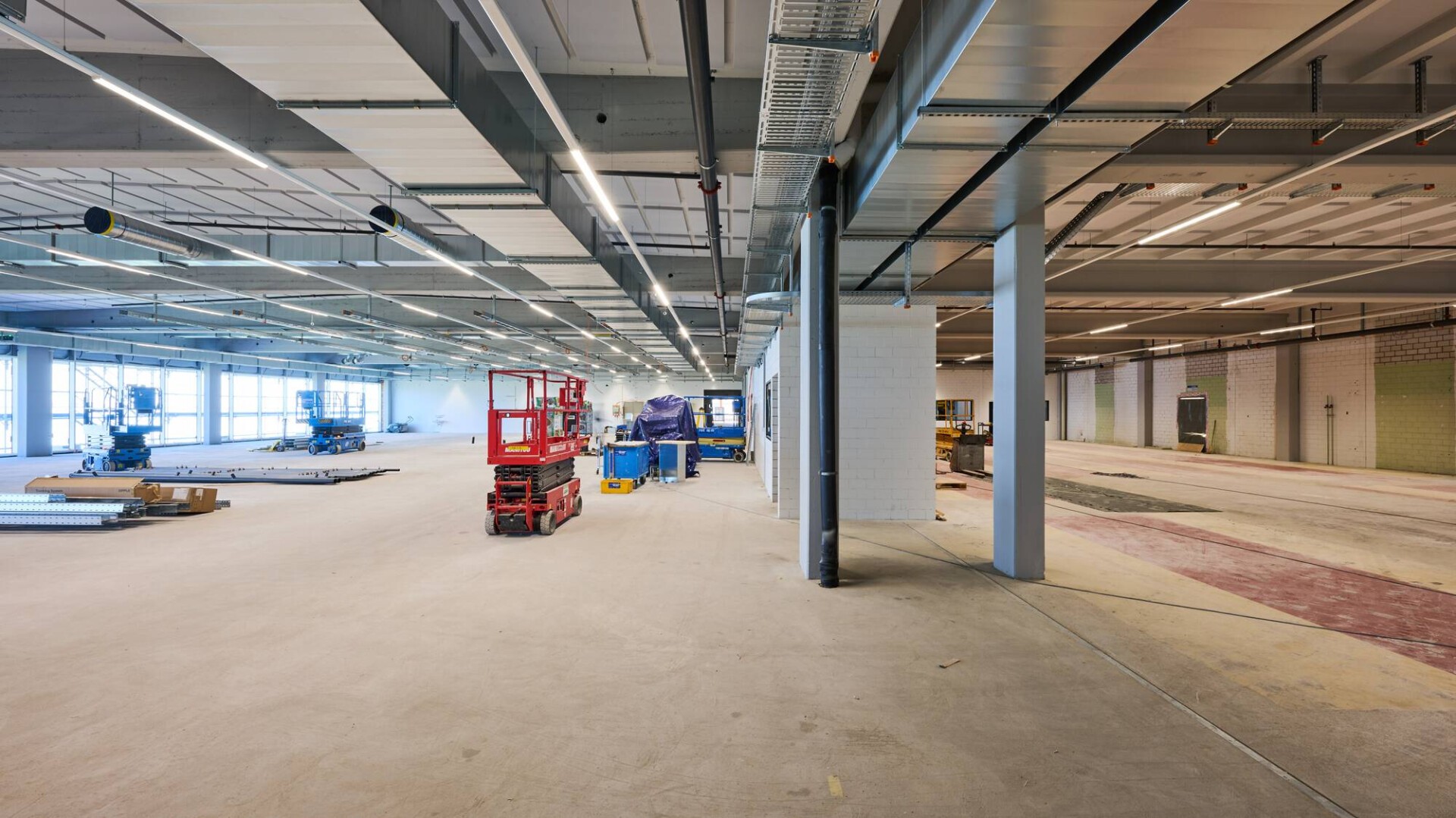 Große, offene Industriefläche im Bau mit freiliegenden Decken, verstreuten Geräten, einer roten Hebebühne, gestapelten Materialien und sichtbaren Arbeitsbereichen auf Betonboden. Helle Beleuchtung und große Fenster an der linken Wand.