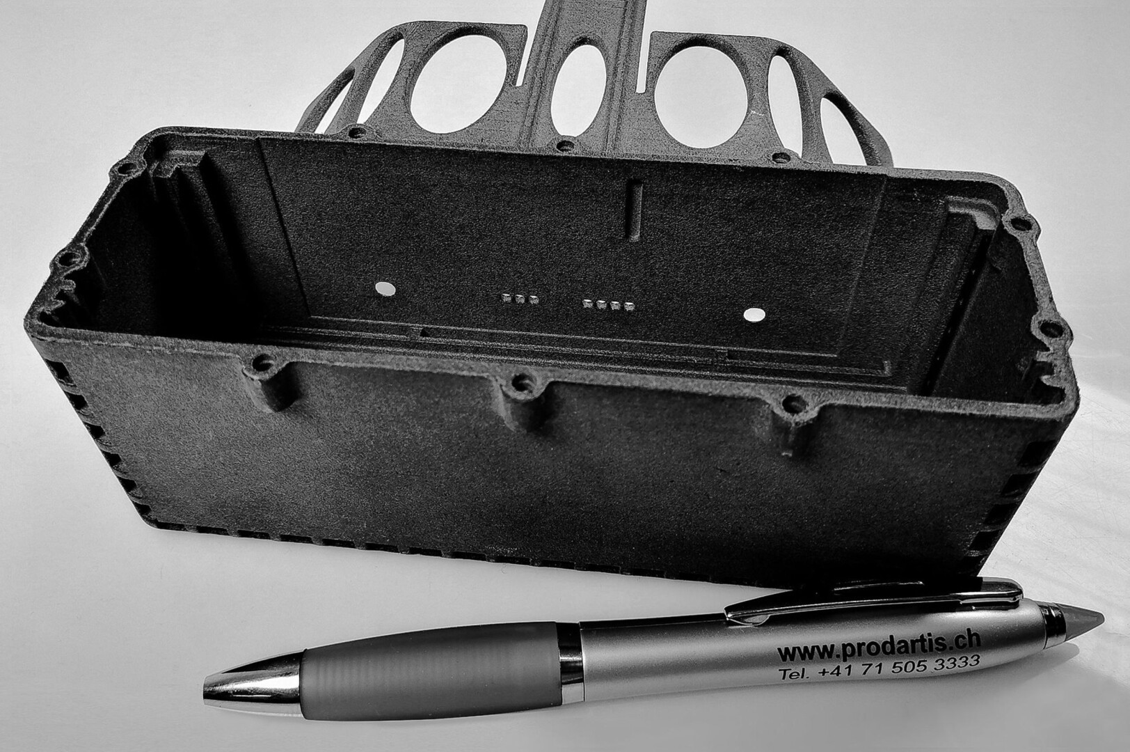 Ein schwarzes rechteckiges Kunststoffgehäuse mit offener Oberseite und abgerundeten Löchern, neben einem metallischen Stift mit der Aufschrift „www.prodaris.ch Tel. +41 71 505 3333“ auf einer weißen Oberfläche.